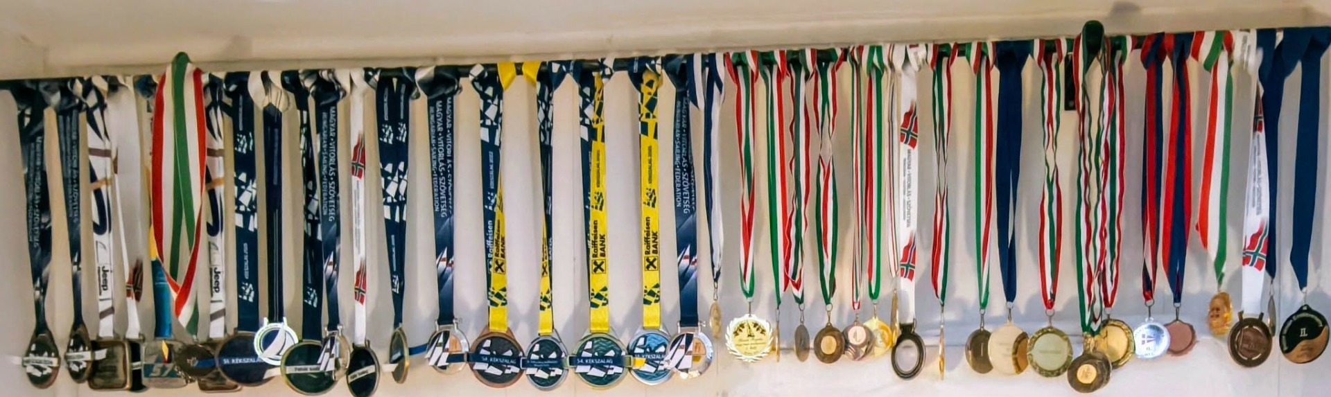 Színes sportérmek és szalagok sorban felakasztva, több nemzeti zászló mintával, háttérben kék-fehér zászló. Kékszalam első hely, vitorlás verseny, vitorlázó kapitány